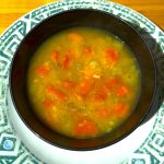 A bowl of split pea soup. The image shows a black bowl with a yellow-brown broth soup with split peas and onions and orange carrots visible.